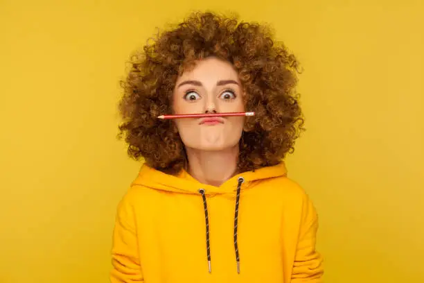 Photo of Closeup of comical woman in urban style hoodie holding pencil with her lips, pretending to have mustache