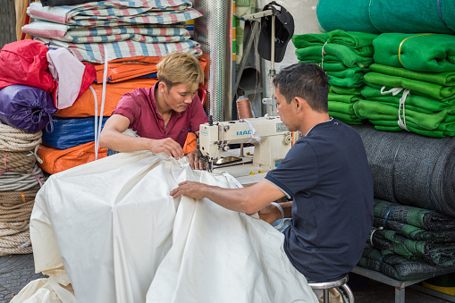 Ho Chi Minh City, Vietnam - February 11, 2019: Vietnamese men stitch a fabric with a sewing machine in the street near their small shop. Saigon is abundant with such small street businesses.
