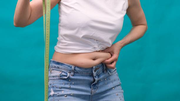 Woman holds a fold of skin on a thick belly, fat female body on a green background. Bold female waist. The concept of weight loss and diet Woman holds a fold of skin on a thick belly, fat female body on a green background. Bold female waist. The concept of weight loss and diet pot belly stock pictures, royalty-free photos & images