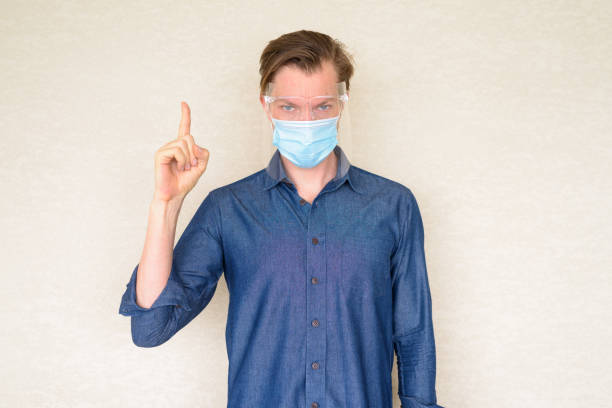 istock Young man with mask and face shield pointing up against concrete wall 1249990341