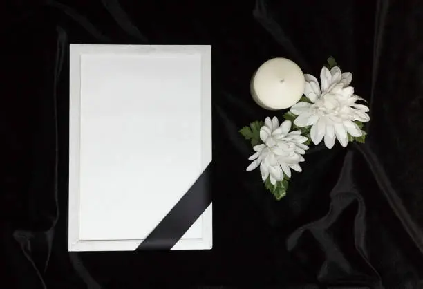 Photo of Condolence card. Memorial frame with black ribbon. White flowers and a burning candle. Black background.