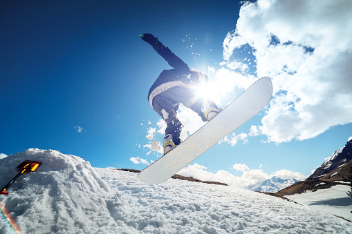Stylish young girl snowboarder does the trick in jumping from a snow kicker against sun the blue sky clouds and mountains in the spring. The concept of spring snowboarding in the mountains and the closing of the winter ski season.