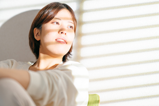 A young woman is sitting on a chair and resting while her face is lit by bright day sunlight with shadow shapes of blinds at home.