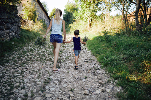 Mother and son with flower bouquets walking on a countryside village road and holding hands.