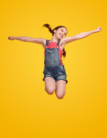 Happy girl in casual clothes smiling and looking away while leaping up with outstretched arms against yellow background