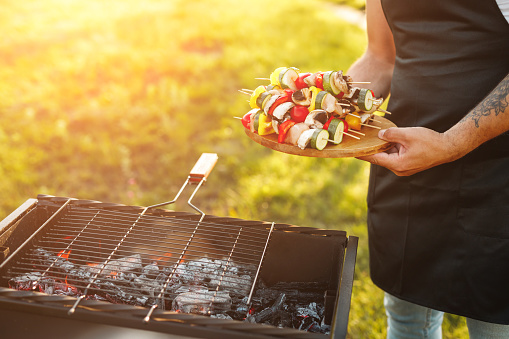 Unrecognizable male with delicious shish kebabs on skewers standing near grill with burning charcoal during picnic in countryside