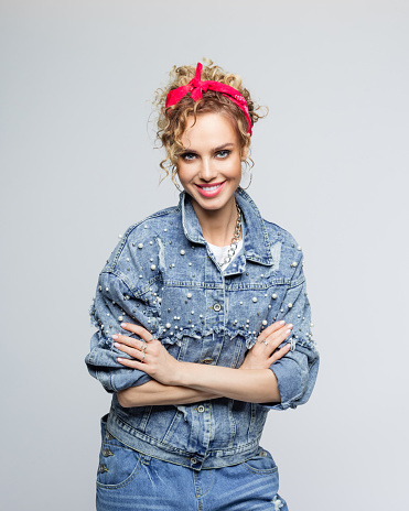 Portrait of blond curly hair confident young woman wearing white t-shirt, denim jacket and red bandana, standing with arms crossed and smiling at camera. Studio shot on grey background.