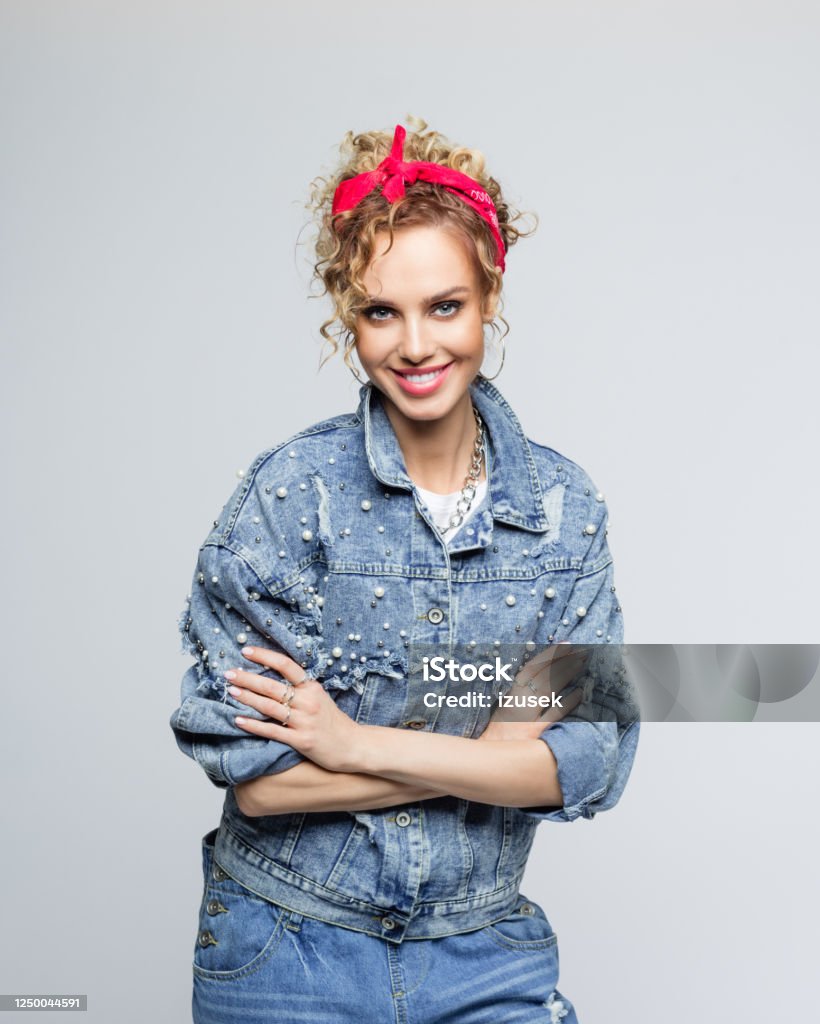 Cheerful young woman in 80's style outfit Portrait of blond curly hair confident young woman wearing white t-shirt, denim jacket and red bandana, standing with arms crossed and smiling at camera. Studio shot on grey background. Arms Crossed Stock Photo