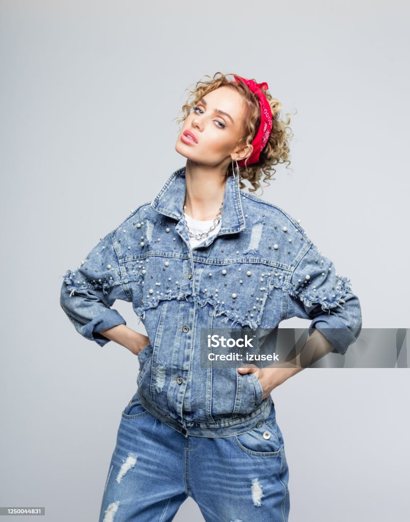 Confident young woman in 80's style outfit Fashion portrait of blond curly hair confident young woman wearing white t-shirt, denim jacket and red bandana, standing with hands in pockets and looking at camera. Studio shot on grey background. 1980-1989 Stock Photo