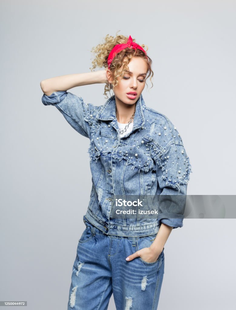 Fashion portrait of young woman in 80's style outfit Blond curly hair confident young woman wearing white t-shirt, denim jacket and pants and red bandana, standing with raised hand and looking away. Studio shot on grey background. 1980-1989 Stock Photo