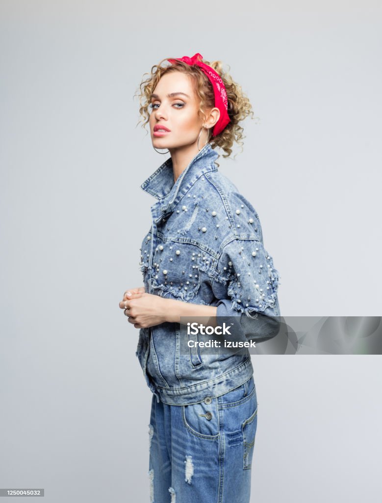 Fashion portrait of young woman in 80's style outfit Blond curly hair confident young woman wearing white t-shirt, denim jacket and pants and red bandana, looking at camera. Studio shot on grey background. 1980-1989 Stock Photo
