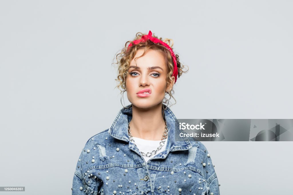 Portrait of disgusted young woman in 80's style outfit Portrait of blond curly hair  young woman wearing white t-shirt, denim jacket and red bandana, grimacing at camera. Studio shot on grey background. 1980-1989 Stock Photo