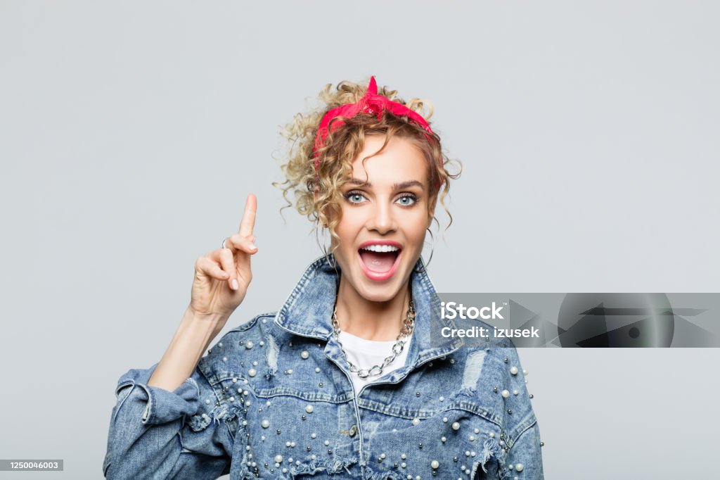 Portrait of excited young woman in 80's style outfit Portrait of blond curly hair confident young woman wearing white t-shirt, denim jacket and red bandana, pointing at copy space and laughing at camera. Studio shot on grey background. 1980-1989 Stock Photo