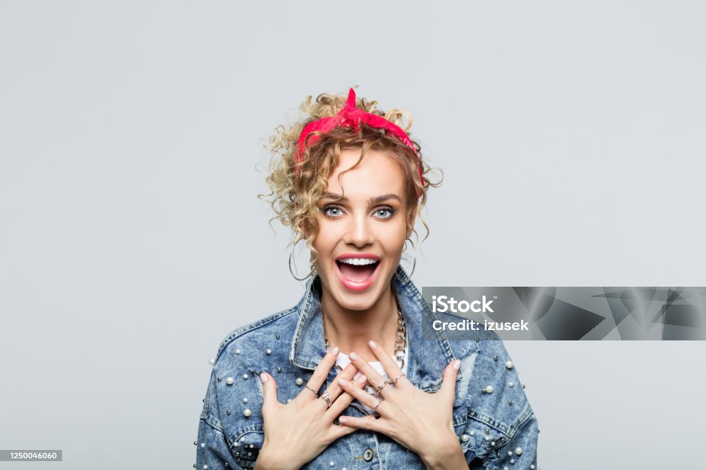 Portrait of surprised young woman in 80's style outfit Portrait of blond curly hair excited young woman wearing white t-shirt, denim jacket and red bandana, laughing at camera. Studio shot on grey background. 1980-1989 Stock Photo