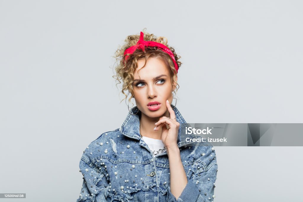 Portrait of thoughtful young woman in 80's style outfit Portrait of blond curly hair young woman wearing white t-shirt, denim jacket and red bandana, looking away with hand on chin. Studio shot on grey background. Choice Stock Photo