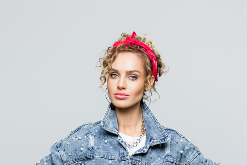 Portrait of blond curly hair confident young woman wearing white t-shirt, denim jacket and red bandana, looking at camera. Studio shot on grey background.