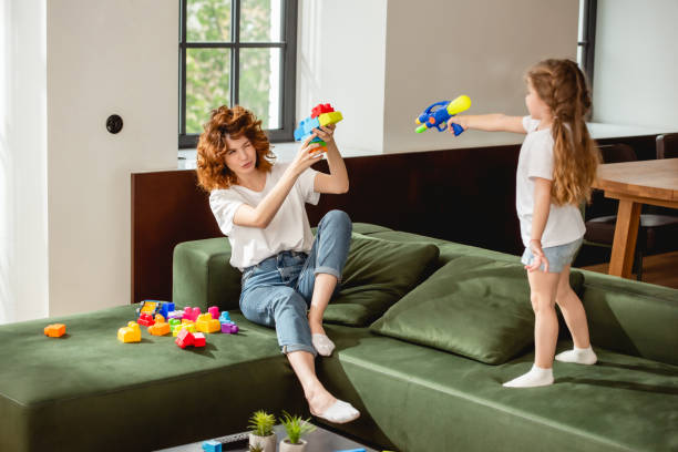 カーリー母ウィンクアイとリビングルームで水銃を持つ娘と遊ぶ - toy gun ストックフォトと画像