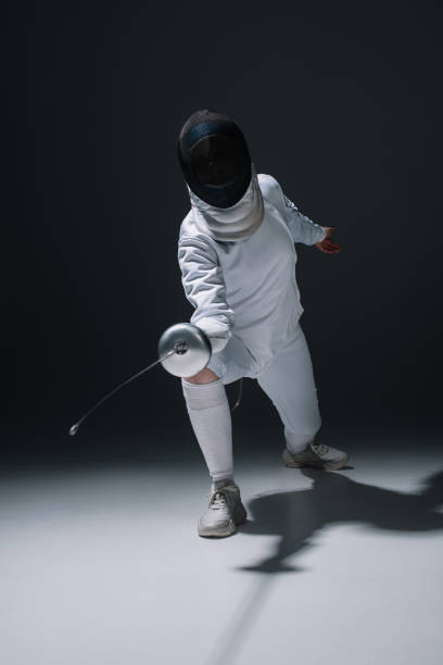 istock Fencer in fencing mask holding rapier while training on white surface on black background 1250046688