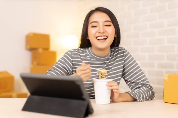 Asia freelance business woman eating instant noodles while working on laptop in living room at home office at night. young Asian girl sitting on desk work overtime, enjoy relax time.female working hard. mujer de negocios independiente de asia comiendo fideos instantáneos mientras trabaja en computadora portátil en la sala de estar en la oficina en casa por la noche. joven asiática chica sentada en el escritorio de trabajo horas extras, disfrutar de rel