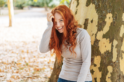 Laughing cute young redhead woman leaning against the trunk of a tree in a high key autumn park peeking out under her long curly hair at the camera