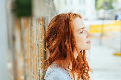 Thoughtful young woman with tousled red hair leaning against an urban wall staring ahead in a close up profile view on a high key background