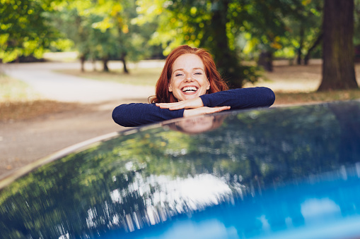 Laughing young woman leaning her chin on a blue car as she looks over the top with a vivacious smile in a wooded park