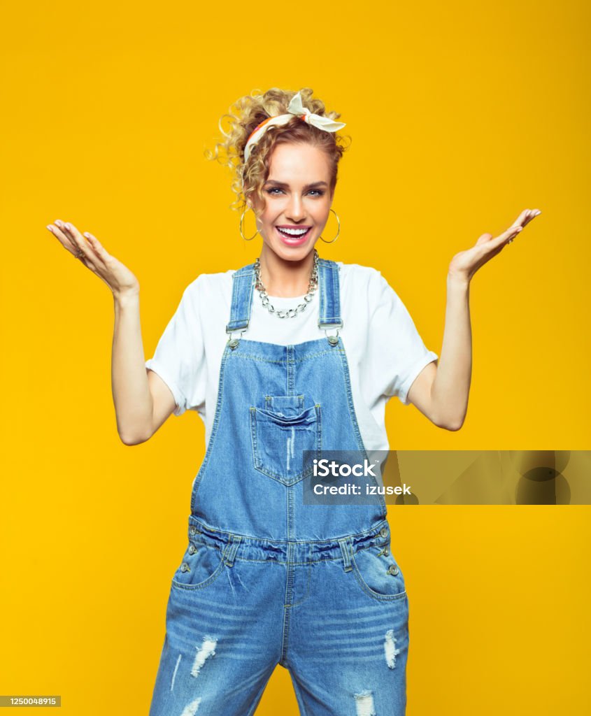Portrait of happy young woman in coveralls on yellow background Portrait of excited young woman wearing white t-shirt, denim dungarees and bandana smiling at camera with raised hands. Studio shot on yellow background. 1980-1989 Stock Photo