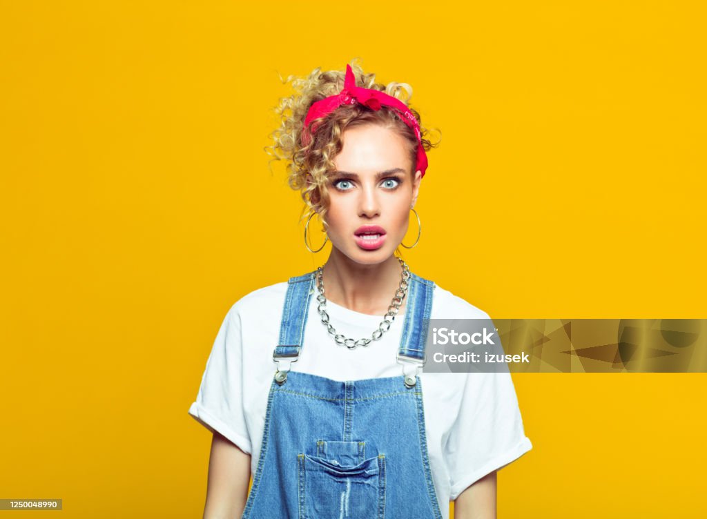 Shocked young woman in 80's style outfit, portrait on yellow background Portrait of surprised young woman wearing white t-shirt, denim dungarees and red bandana staring at camera. Studio shot on yellow background. Displeased Stock Photo