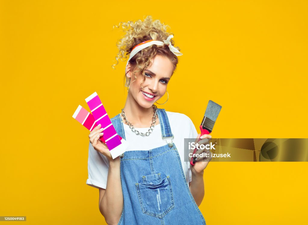 Young woman in coveralls holding color swatch and paintbrush Portrait of cheerful young woman wearing white t-shirt, denim dungarees and bandana holding color swatch and paintbrush. Studio shot on yellow background. Colored Background Stock Photo