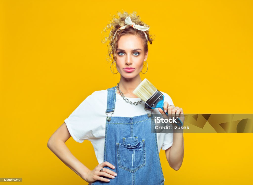 Young woman in coveralls holding paintbrush Portrait of confident young woman wearing white t-shirt, denim dungarees and bandana holding paintbrush, looking at camera. Studio shot on yellow background. DIY Stock Photo