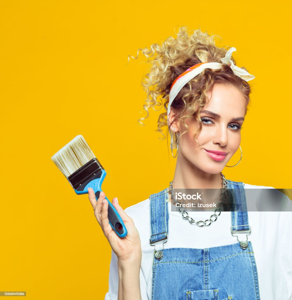 Young woman in coveralls holding paintbrush Portrait of happy young woman wearing white t-shirt, denim dungarees and bandana holding paintbrush, smiling at camera. Studio shot on yellow background. DIY Stock Photo