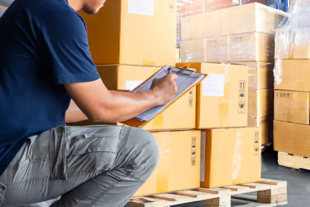 Warehouse worker holding clipboard writing on checklist for delivering shipment goods. Package boxes, Cargo shipment export, Industry warehouse shipping logistics and transport.