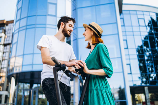 A young romantic couple with electric scooters on a date, walking in the city. Young woman in hat and man enjoy a walk stock photo