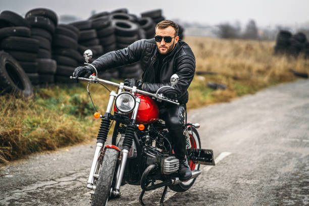 istock Bearded man in sunglasses and leather jacket looking at the camera while sitting on a motorcycle on the road. Behind him is a row of tires 1250072583