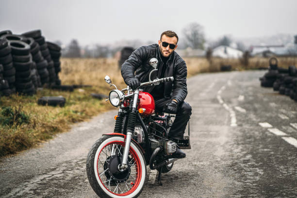 istock Bearded man in sunglasses and leather jacket looking at the camera while sitting on a motorcycle on the road. Behind him is a row of tires 1250072597