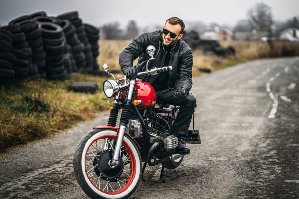 istock Bearded man in sunglasses and leather jacket smiling while sitting on a red motorcycle on the road. Behind him is a row of tires 1250072598