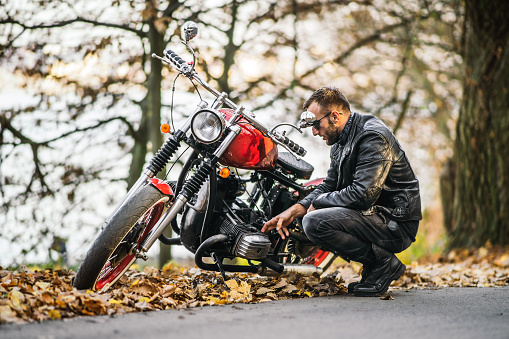 Bearded brutal man in sunglasses and leather jacket sitting on a motorcycle on the road in the forest with blured colorful background.