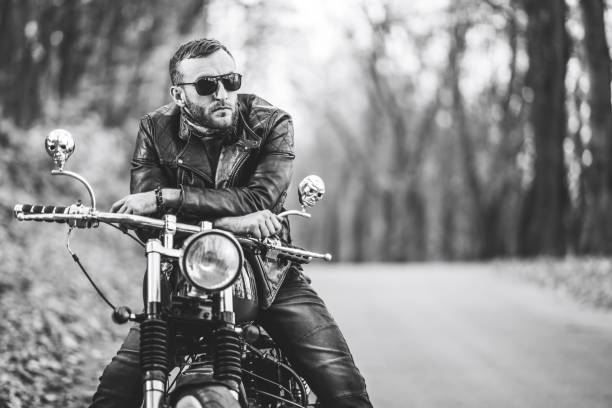 Bearded brutal man in sunglasses and leather jacket sitting on a motorcycle on the road in the forest stock photo