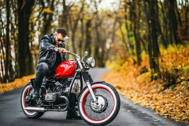 Bearded brutal man in sunglasses and leather jacket sitting on a motorcycle on the road in the forest stock photo
