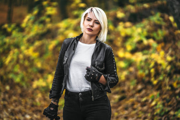 Pretty blonde biker styled women in black leather jacket with sunglasses standing outdoors in the forest stock photo