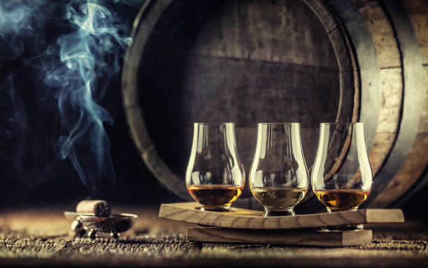 Glencairn whiskey tasting cups on a wooden serving, with a whisky barrel in the dark background and a smoking cigar next to it Glencairn whiskey tasting cups on a wooden serving, with a whisky barrel in the dark background and a smoking cigar next to it. glass of bourbon stock pictures, royalty-free photos & images