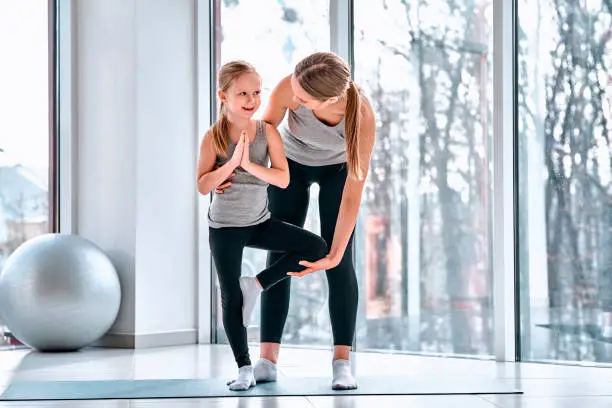 Full length portrait of mother gently teaching little daughter her first yoga notions careful near kids body helping daughter finished action in the fitness class at school.