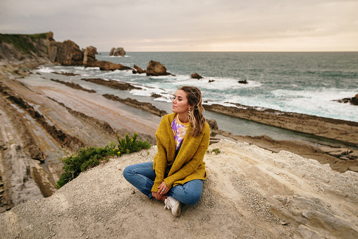 Young woman sitting and relaxing by the sea. She wears a yellow jacket and jeans