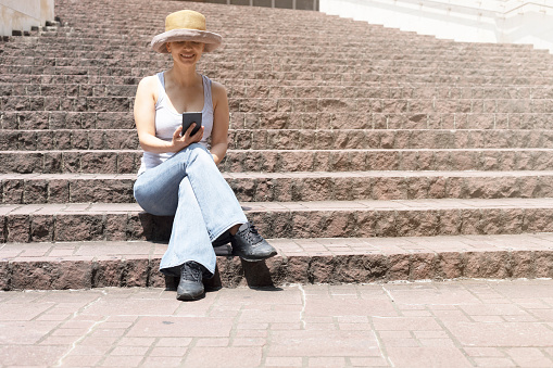 Unrecognizable female wearing a hat is sitting alone on a staircase using her smart phone.