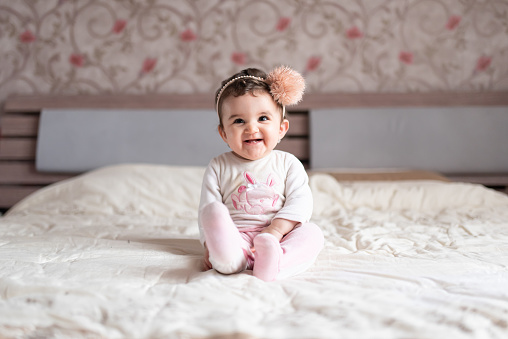 a one year old baby girl sitting on a bed, playing and smiling to the camera.