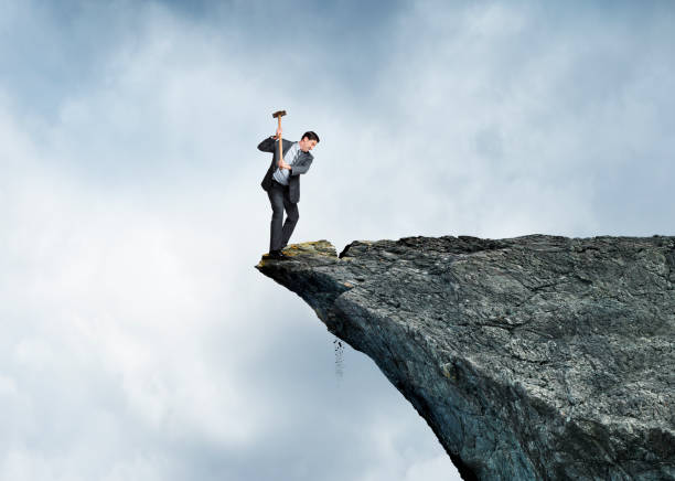 Businessman On Wrong Side A businessman swings a sledge hammer against a rocky cliff as he stands on the wrong side placing himself at great risk. sabotage photos stock pictures, royalty-free photos & images