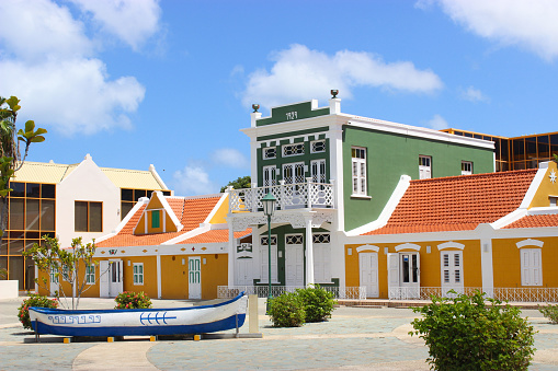 Several colored buildings stand on the street. The sun shines brightly, the sky is blue. In the foreground on the ground is a white-blue boat. Aruba August 2014
