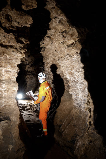 istock Man walking and exploring dark cave with light headlamp and map in his hand underground. Mysterious deep dark, explorer discovering mystery moody tunnel looking on rock wall inside. 1250126145