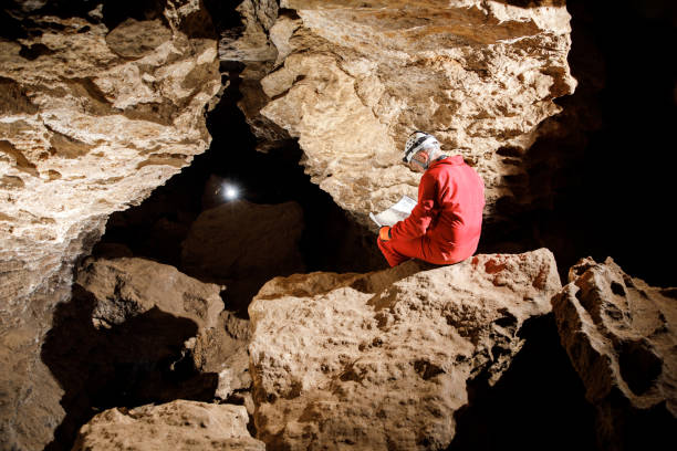 istock Man walking and exploring dark cave with light headlamp underground. Mysterious deep dark, explorer discovering mystery moody tunnel looking on rock wall inside. 1250126546