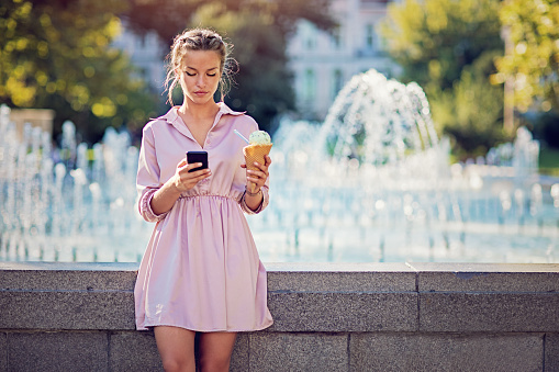 Young woman is eating ice cream and texting at a city fountain